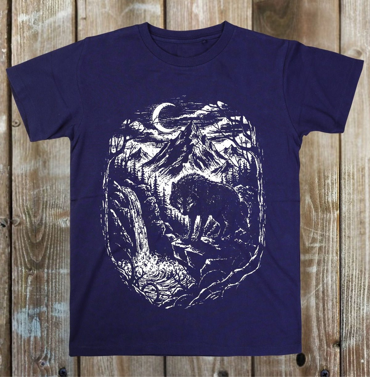 A navy blue short-sleeve crewneck t-shirt with a white distressed graphic print of a wolf, mountains, and a crescent moon, displayed flat against a rustic wooden plank background.