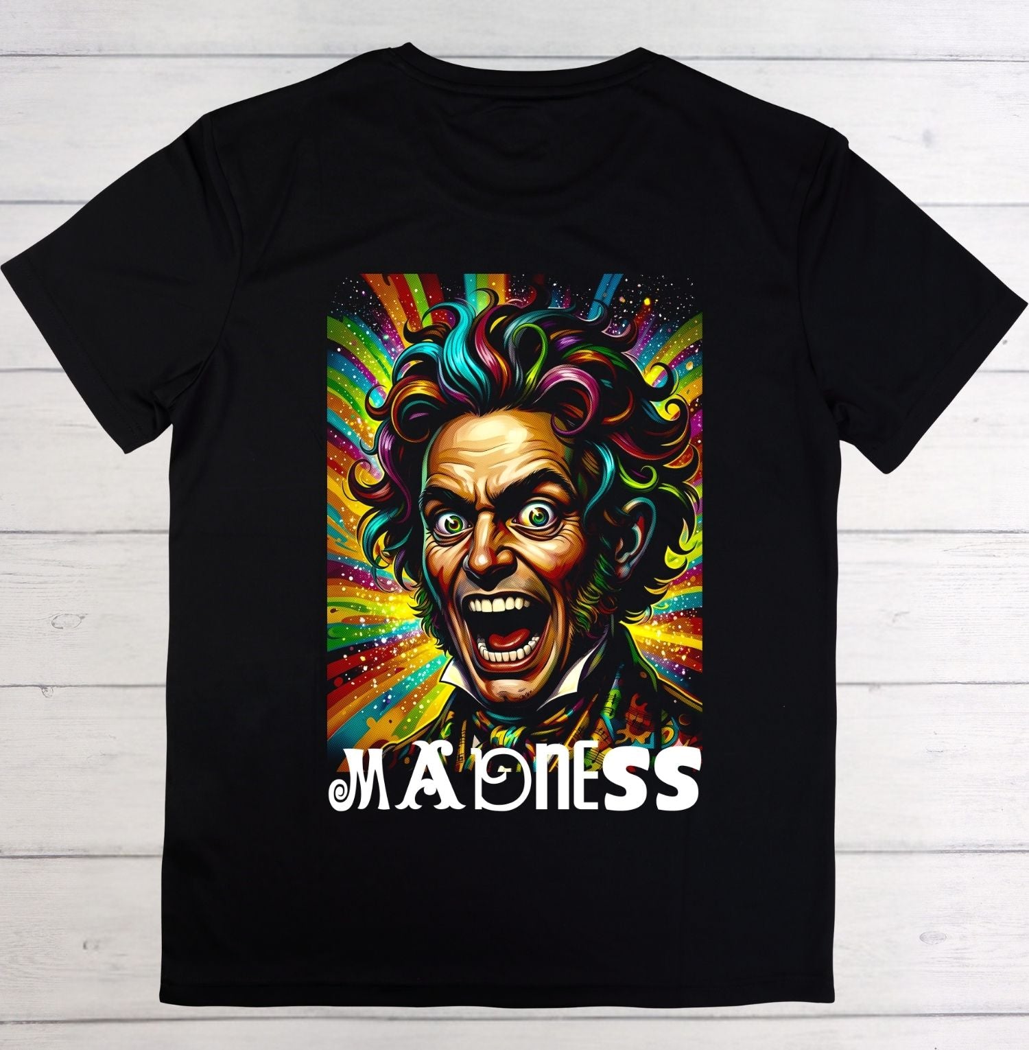 A black short-sleeve crewneck t-shirt laid flat on a white wood plank background, featuring a colorful graphic of a screaming clown face with rainbow-colored hair and the word 'MADENESS' printed below in white.