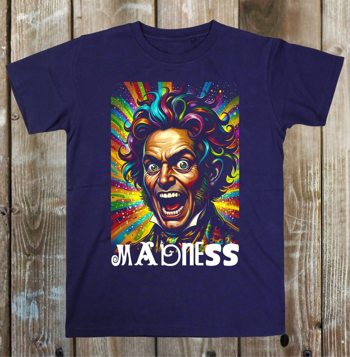 A navy blue short-sleeve t-shirt with a colorful graphic print of a screaming, wild-eyed man with rainbow hair and the word 'MADNESS' in white text below, displayed flat against a rustic wooden plank background.