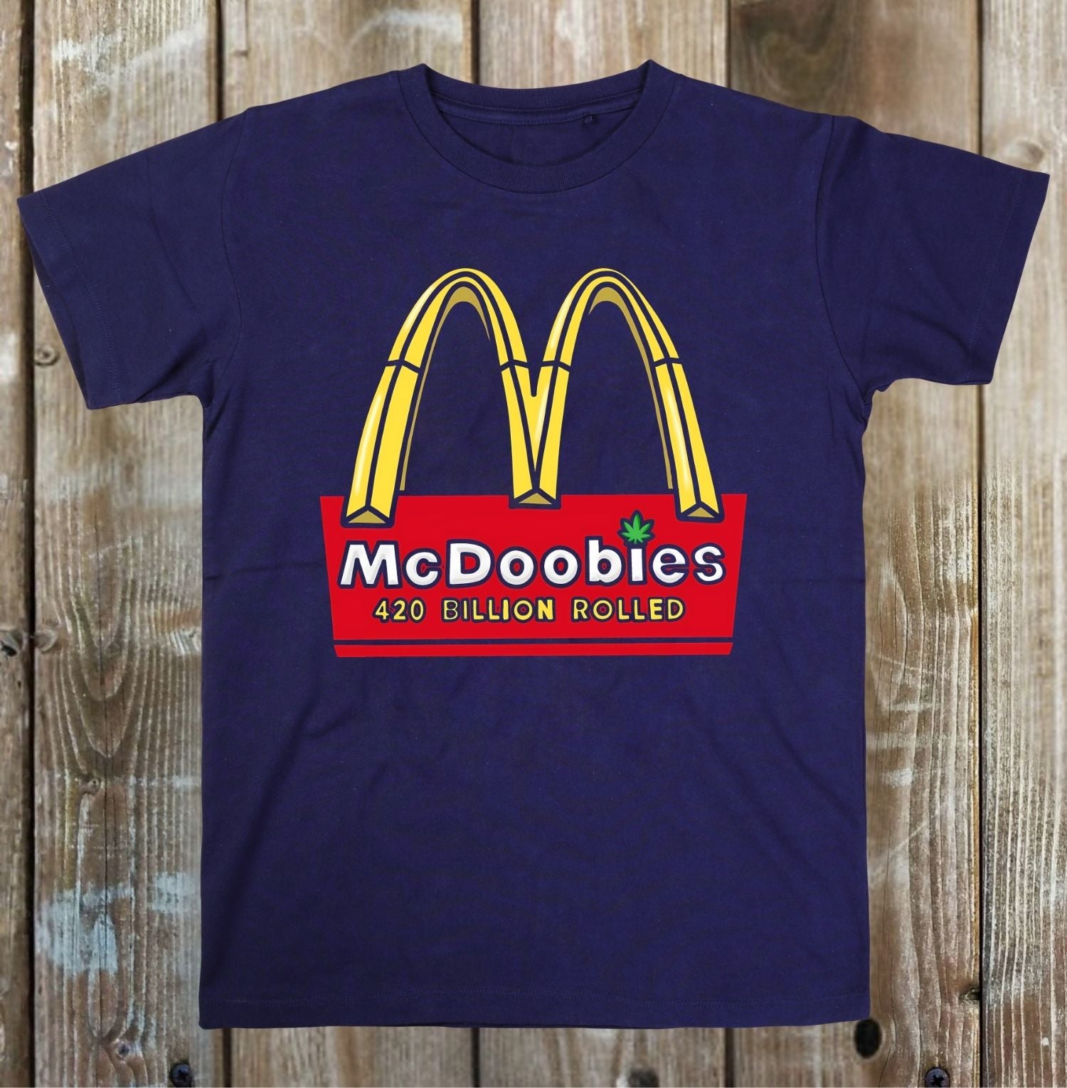 A navy blue t-shirt featuring a parody graphic of the golden arches with 'McDoobies' and '420 BILLION ROLLED' written on a red background, displayed flat on a rustic wooden plank surface.