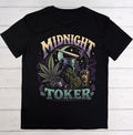 A black short-sleeve crewneck t-shirt laid flat on a white wood plank background. The front features a large graphic print with the words 'MIDNIGHT TOKER' in stylized purple and green lettering, surrounded by illustrations of a cannabis leaf, a rolling machine, a jar, a dropper bottle, and a moon.