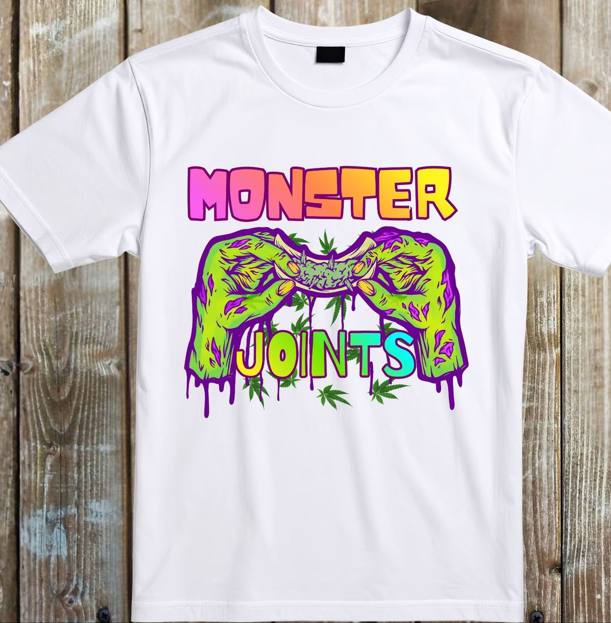 A white short-sleeve crewneck t-shirt laid flat on a rustic wooden plank background. The shirt features a colorful graphic print with the words 'MONSTER JOINTS' in a dripping neon style, with two green hands holding a joint above the text.
