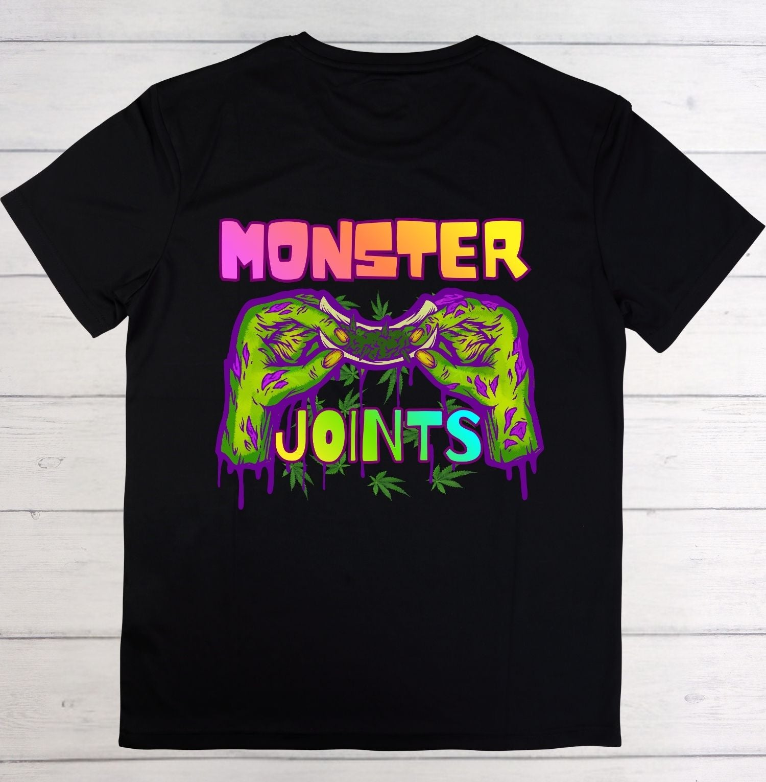 A black short-sleeve t-shirt laid flat on a white wood plank background, featuring a colorful graphic of two green zombie hands holding a beer glass, with the words 'MONSTER JOINTS' in neon pink, yellow, and blue dripping text.