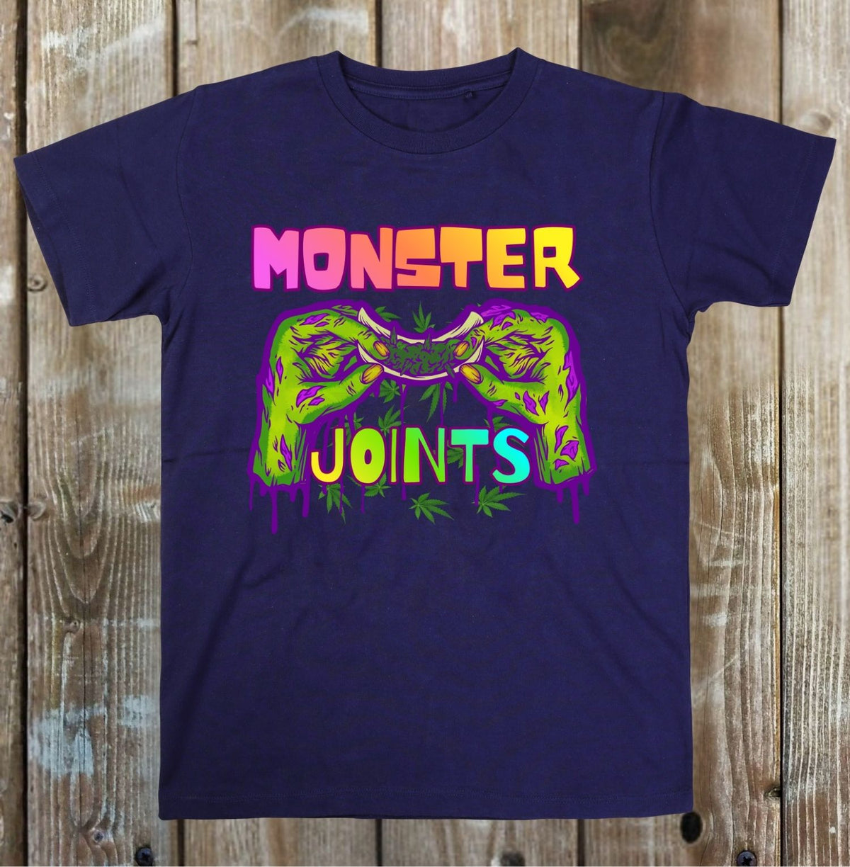 A navy blue short-sleeve t-shirt with a colorful graphic on a rustic wooden plank background. The graphic features the text 'MONSTER JOINTS' in a dripping neon style, with a green joint illustration in the center.