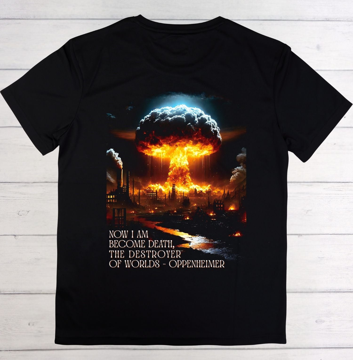 A black short-sleeve t-shirt laid flat on a light wood background, featuring a large graphic print of a nuclear mushroom cloud over a burning industrial landscape, with the text 'NOW I AM BECOME DEATH, THE DESTROYER OF WORLDS - OPPENHEIMER' printed below.