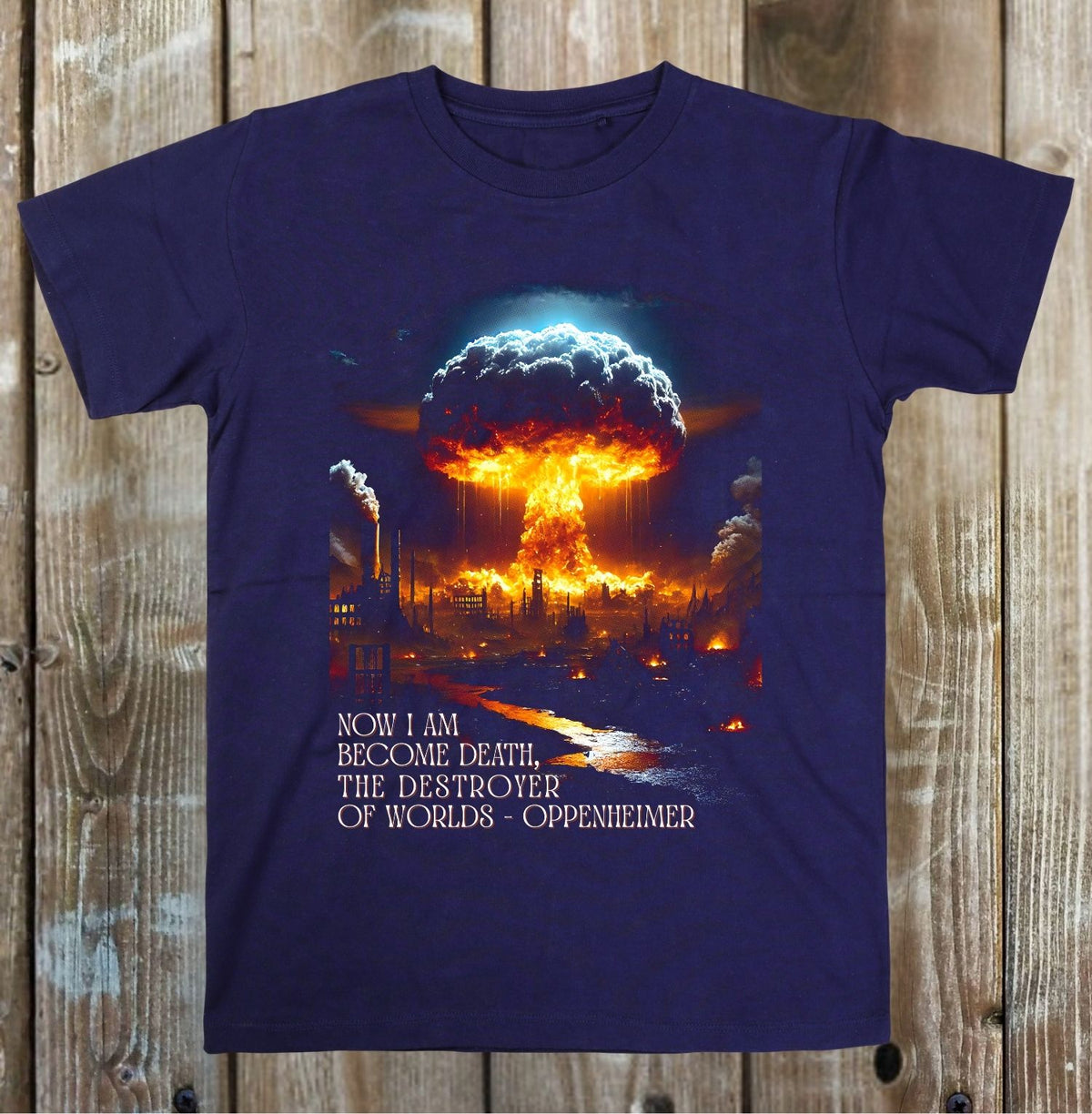 A navy blue short-sleeve t-shirt with a graphic of a nuclear mushroom cloud over a burning industrial landscape, with the text 'NOW I AM BECOME DEATH, THE DESTROYER OF WORLDS - OPPENHEIMER' printed below, displayed flat on a rustic wooden plank background.