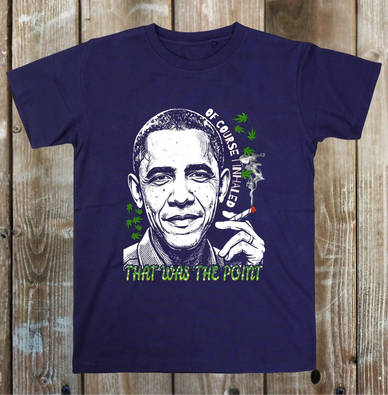 A navy blue short-sleeve t-shirt with a graphic print of Barack Obama smoking a joint, surrounded by green cannabis leaves and the text 'OF COSE ENDEARED' and 'THAT WAS THE POINT'. The t-shirt is displayed flat against a rustic wooden plank background.