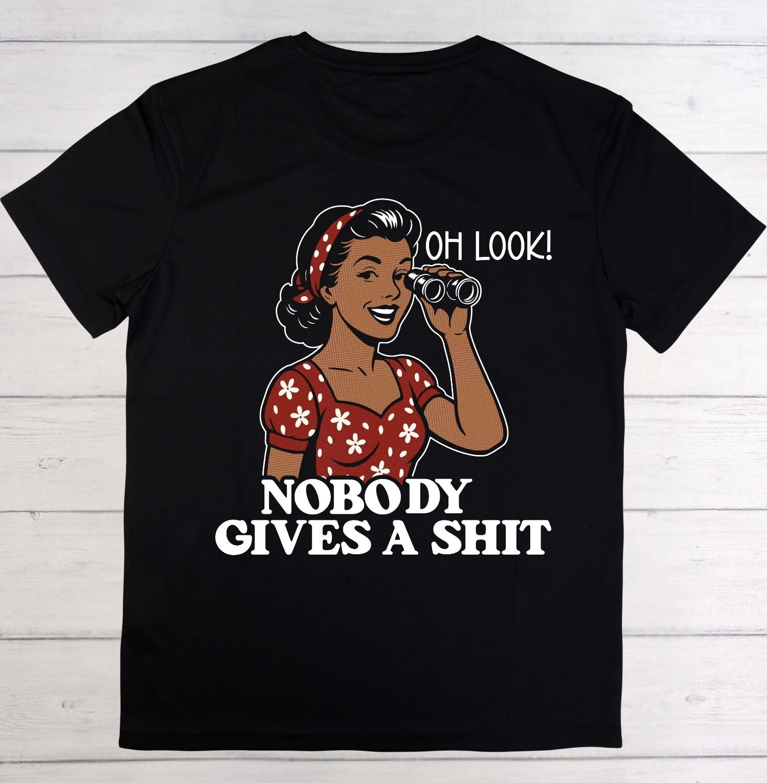 A black short-sleeve t-shirt laid flat on a white wood background, featuring a retro-style graphic of a woman in a red and white floral dress holding binoculars, with the text 'OH LOOK! NOBODY GIVES A SHIT' in white.