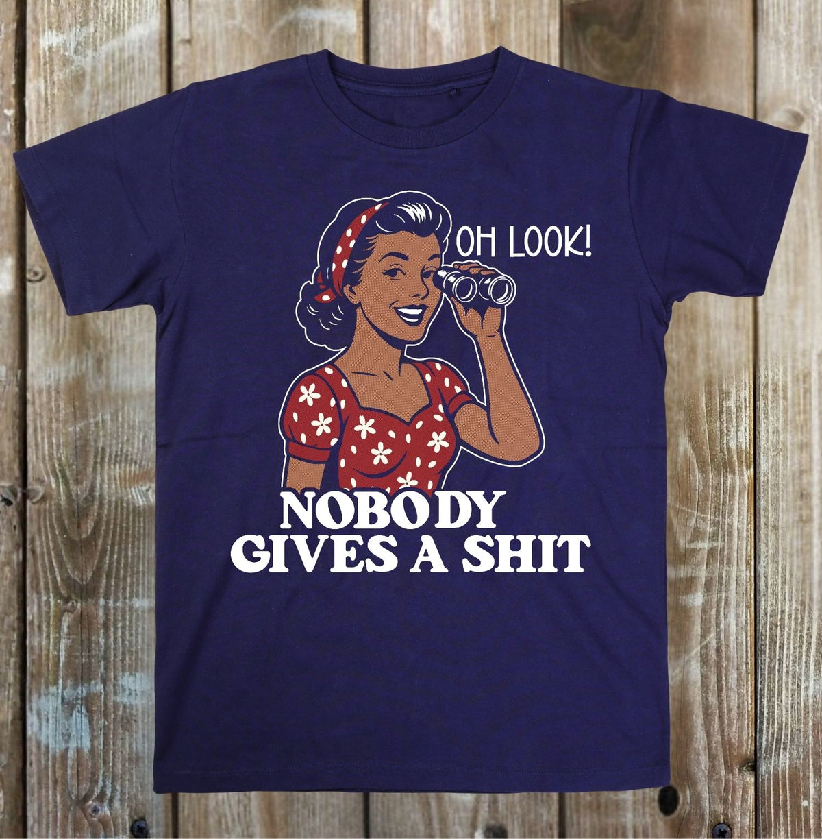 A navy blue short-sleeve t-shirt with a retro-style graphic of a woman in a red floral dress looking through binoculars, accompanied by the text 'OH LOOK! NOBODY GIVES A SHIT' in white, displayed flat against a rustic wooden plank background.