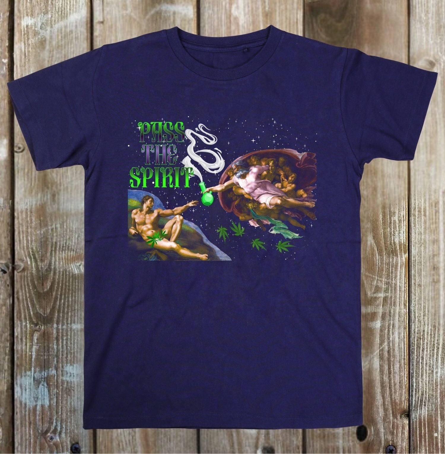 A navy blue short-sleeve t-shirt with a colorful graphic print on the front, laid flat on a rustic wooden plank background. The graphic features the text 'Give Me The Spirit' in green and white, alongside a classical painting of a figure in a shell with marijuana leaves.
