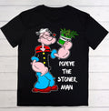 A black short-sleeve t-shirt laid flat on a white wood plank background, featuring a large graphic of Popeye the Sailor Man holding a green cannabis plant, with the text 'POPEYE THE STONER MAN' printed in white.