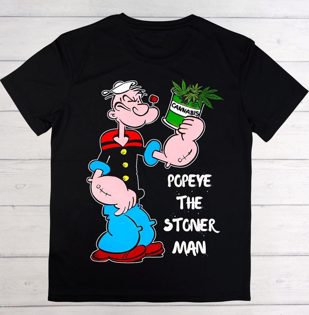 A black short-sleeve t-shirt laid flat on a white wood plank background, featuring a large graphic of Popeye the Sailor Man holding a green cannabis plant, with the text 'POPEYE THE STONER MAN' printed in white.