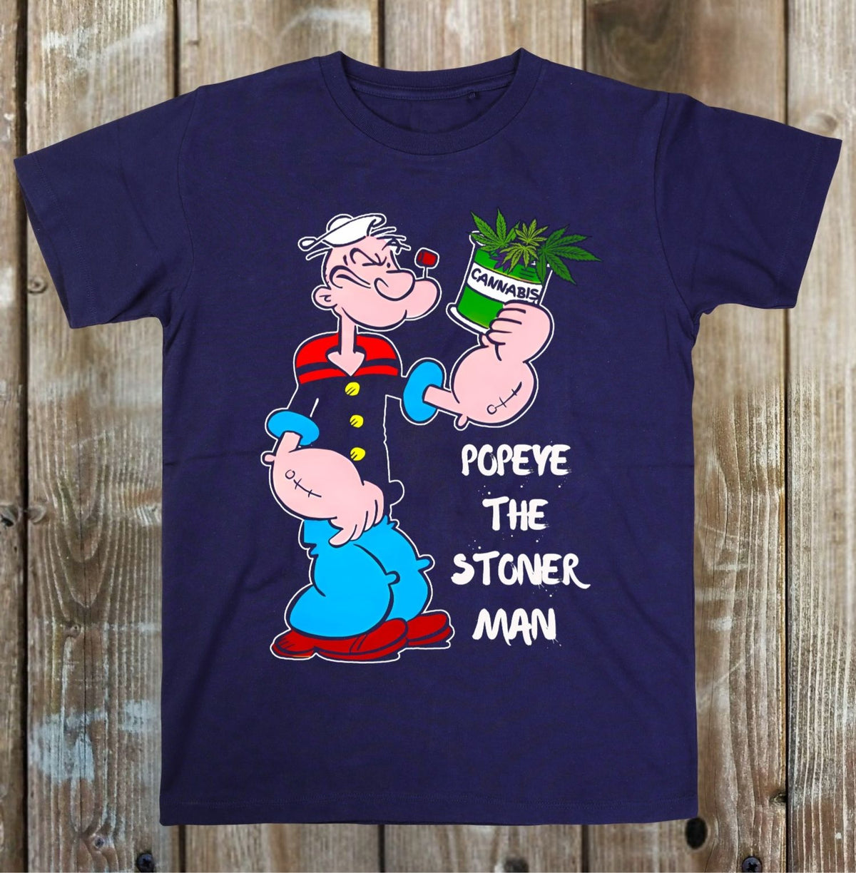A navy blue short-sleeve t-shirt with a cartoon graphic of Popeye holding a green can labeled 'CANNABIS' next to the text 'POPEYE THE STONER MAN', displayed flat against a rustic wooden plank background.