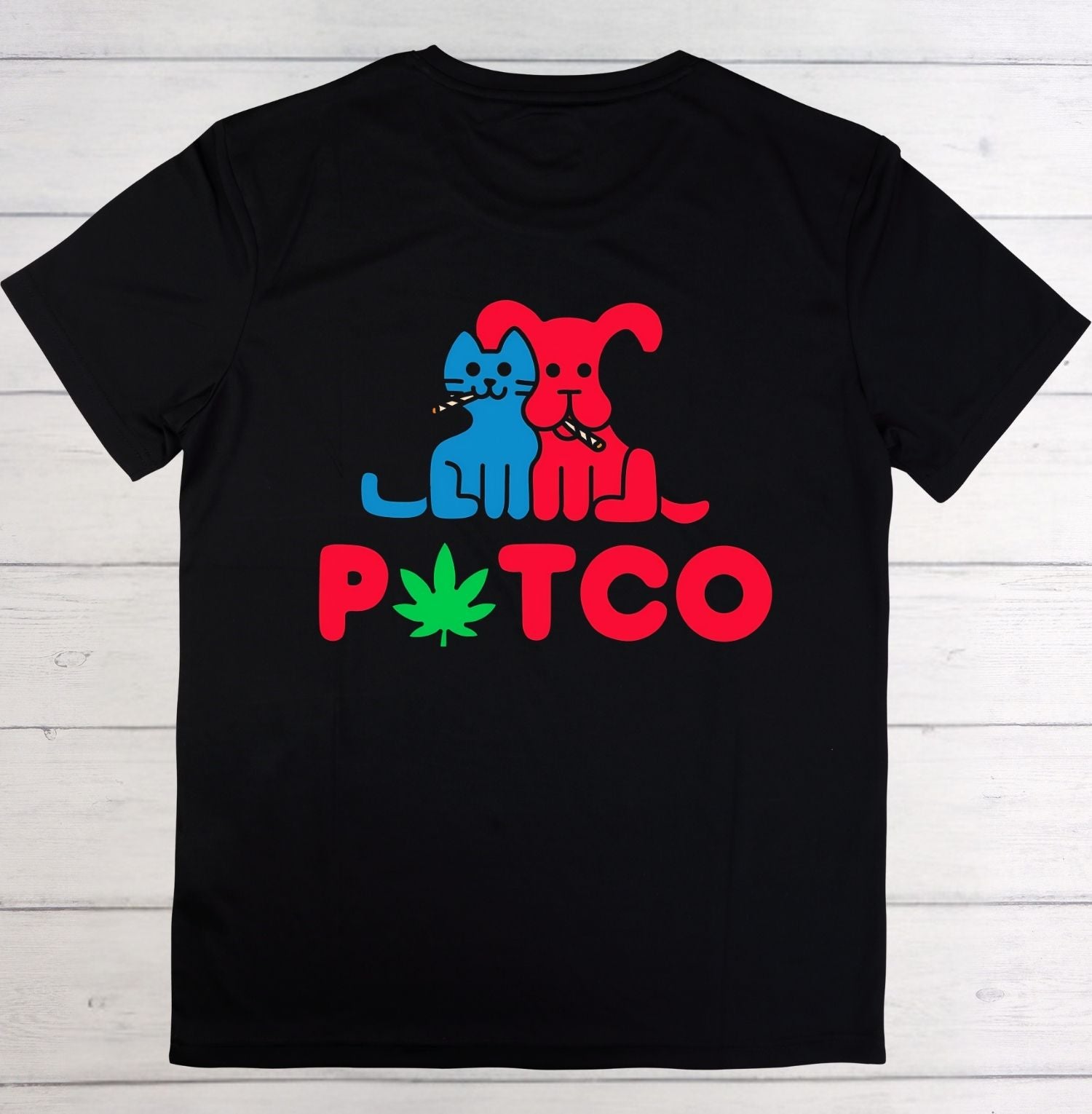 A black short-sleeve t-shirt laid flat on a white wood plank background, featuring a colorful graphic of a blue cat and a red dog smoking, with the word 'PTCO' in red below and a green cannabis leaf replacing the 'O'.
