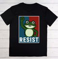 A black short-sleeve t-shirt laid flat on a white wood plank background, featuring a graphic print of a cartoon green frog raising its fist in front of a blue and red split background with the word 'RESIST' in white block letters at the bottom.