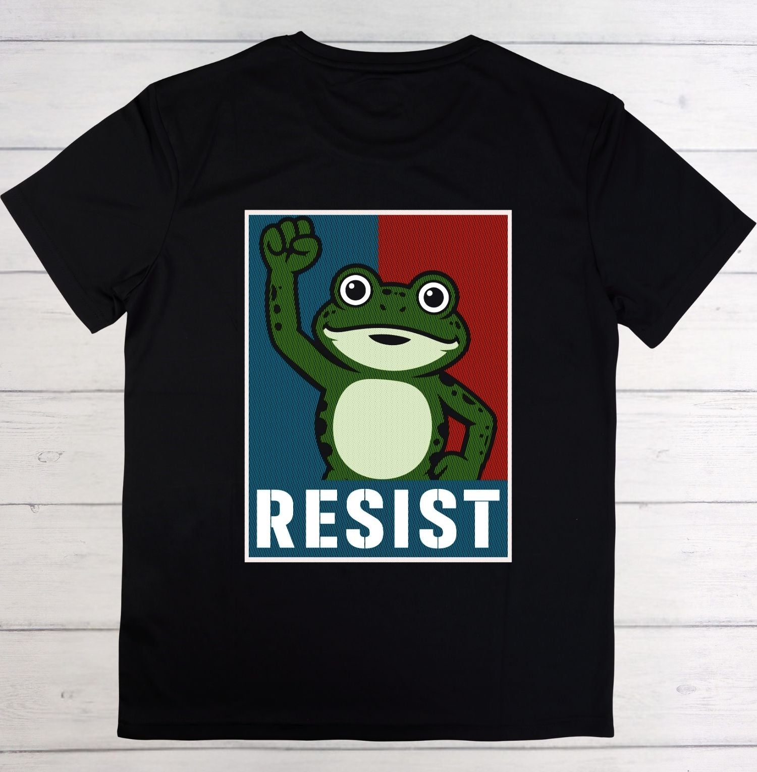 A black short-sleeve t-shirt laid flat on a white wood plank background, featuring a graphic print of a cartoon green frog raising its fist in front of a blue and red split background with the word 'RESIST' in white block letters at the bottom.