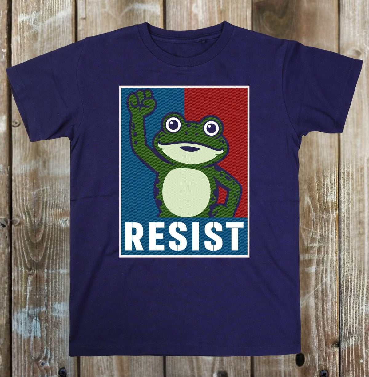 A navy blue short-sleeve t-shirt with a graphic print of a green frog raising its fist against a blue and red background with the word 'RESIST' in white. The t-shirt is displayed flat on a rustic wooden plank background.