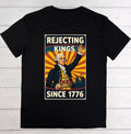 A black short-sleeve t-shirt laid flat on a white wood plank background, featuring a large vintage-style graphic print on the front with the text 'REJECTING KINGS SINCE 1776' and an illustration of George Washington.