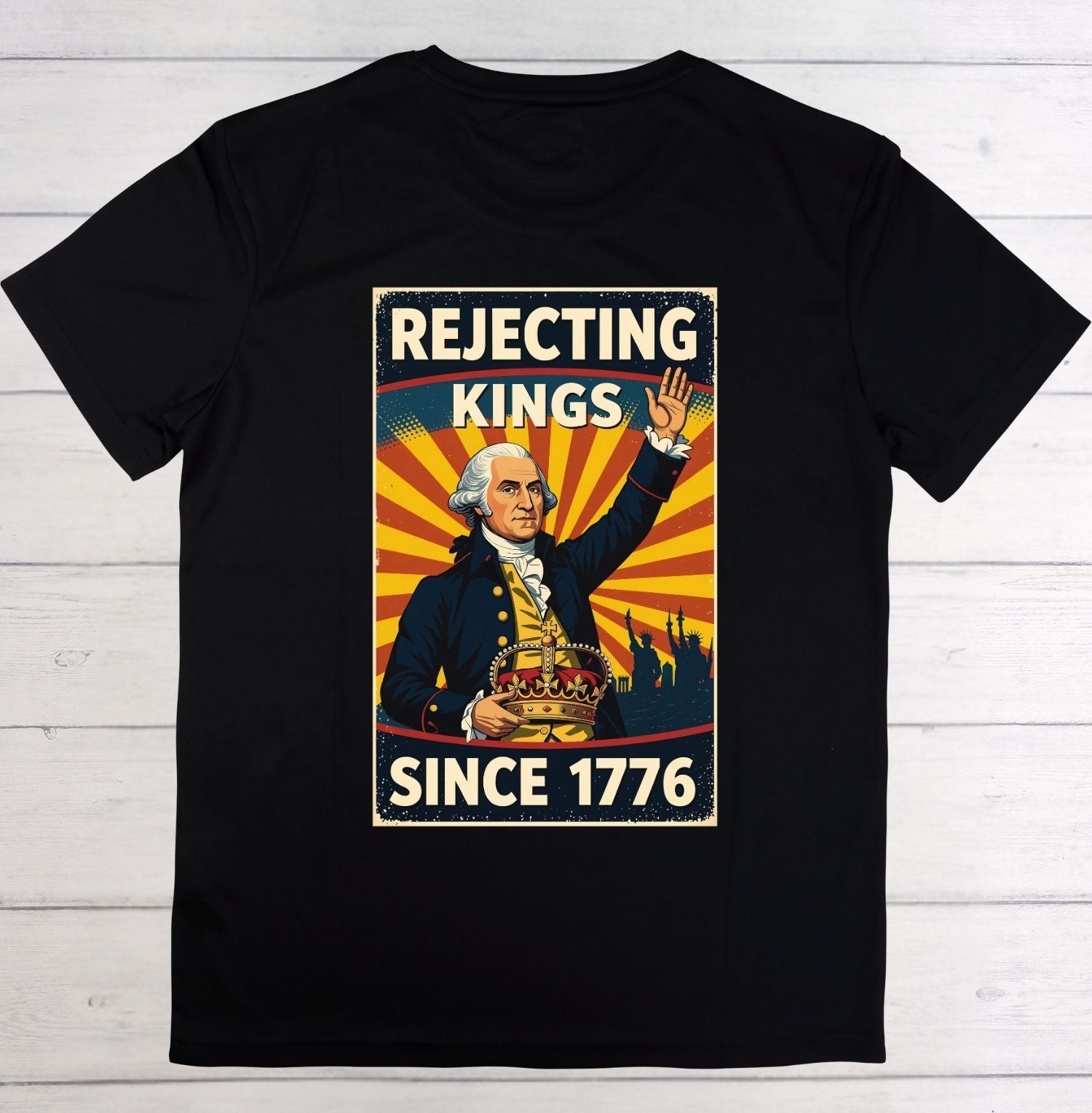 A black short-sleeve t-shirt laid flat on a white wood plank background, featuring a large vintage-style graphic print on the front with the text 'REJECTING KINGS SINCE 1776' and an illustration of George Washington.