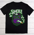A black short-sleeve t-shirt laid flat on a white wood plank background, featuring a large graphic of a purple gorilla smoking with green smoke and the word 'SMOKE' in green and white.