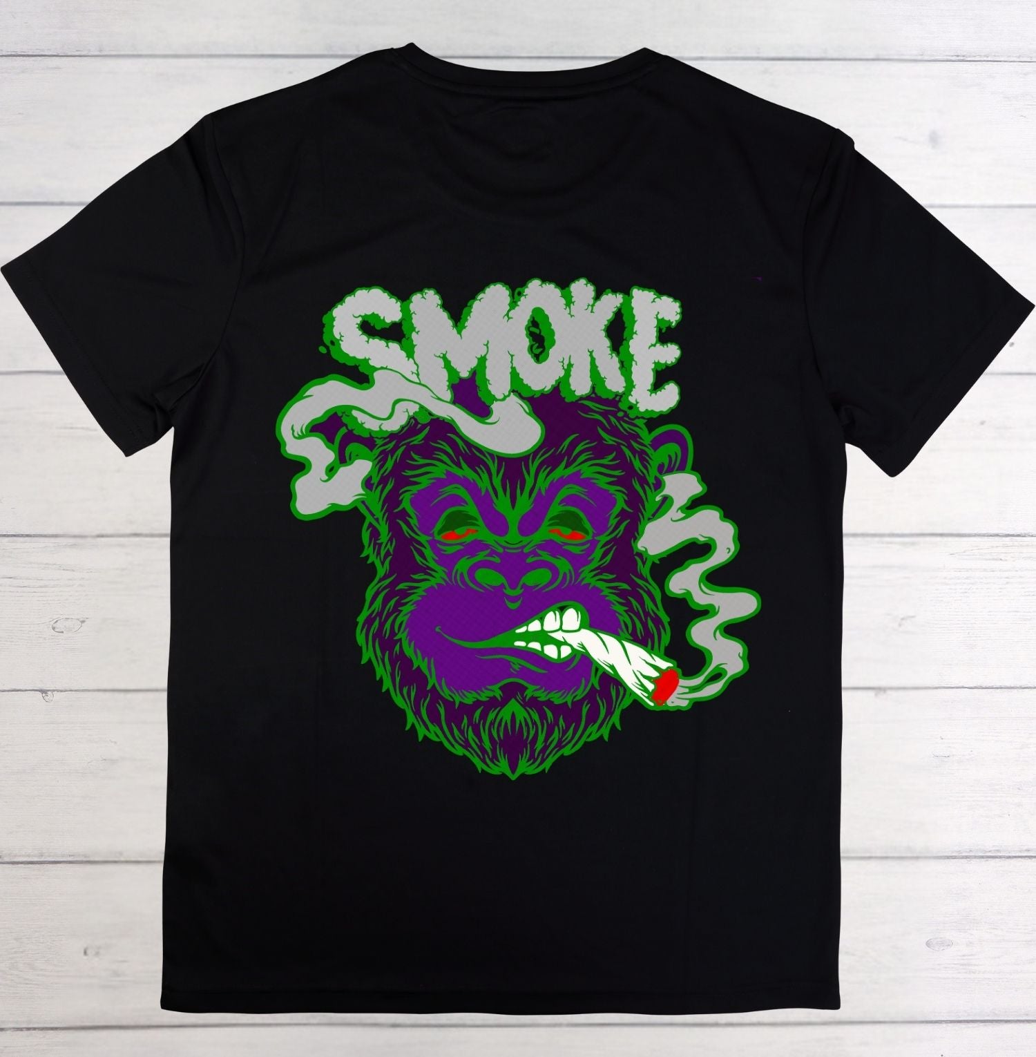 A black short-sleeve t-shirt laid flat on a white wood plank background, featuring a large graphic of a purple gorilla smoking with green smoke and the word 'SMOKE' in green and white.