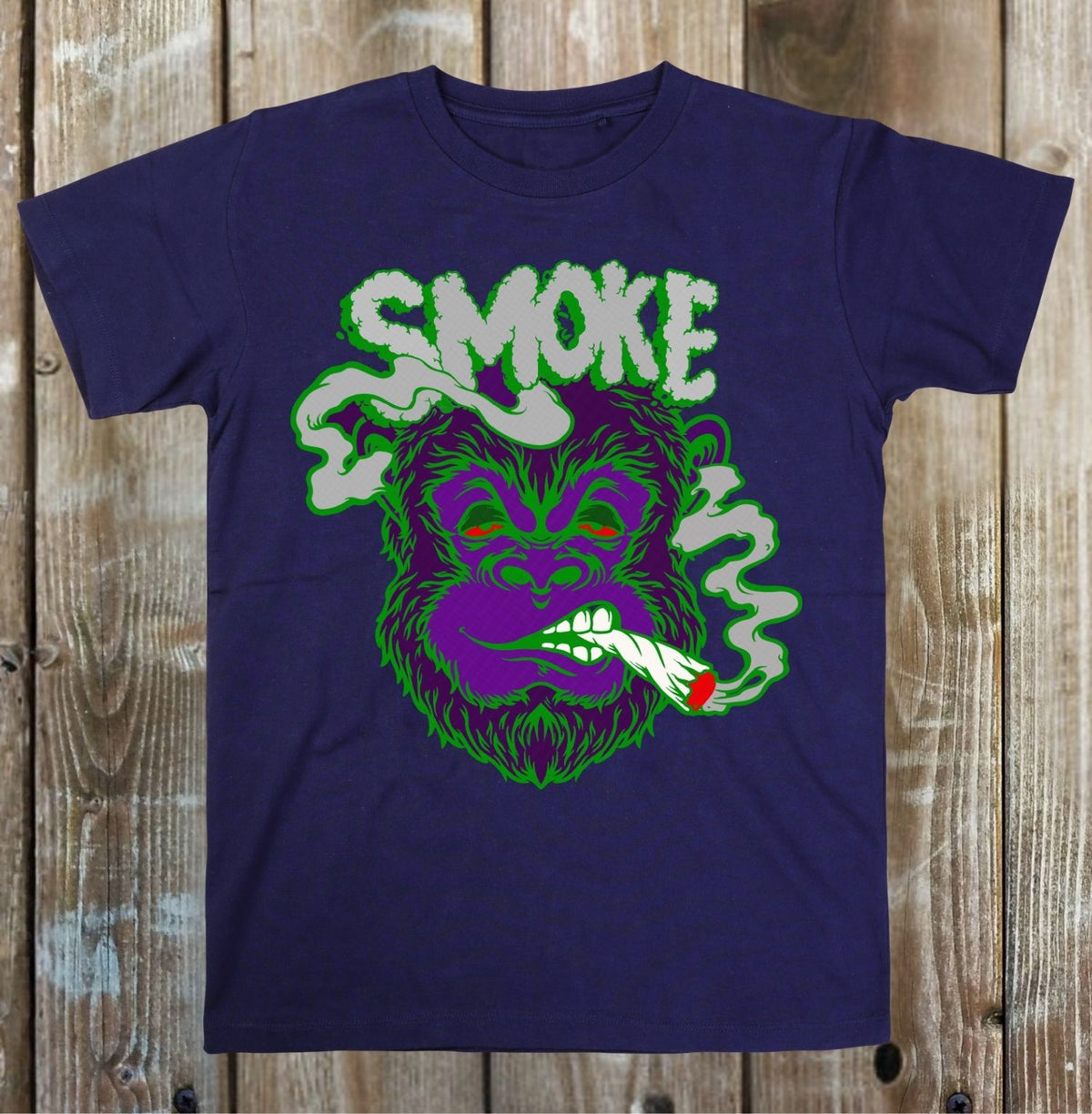 A navy blue short-sleeve t-shirt with a graphic of a purple gorilla smoking, with the word 'SMOKE' in green and white smoke above it, displayed flat against a rustic wooden plank background.