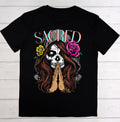 Black t-shirt with a graphic design of a skull with praying hands and flowers, labeled 'SACRED', on a wooden background.