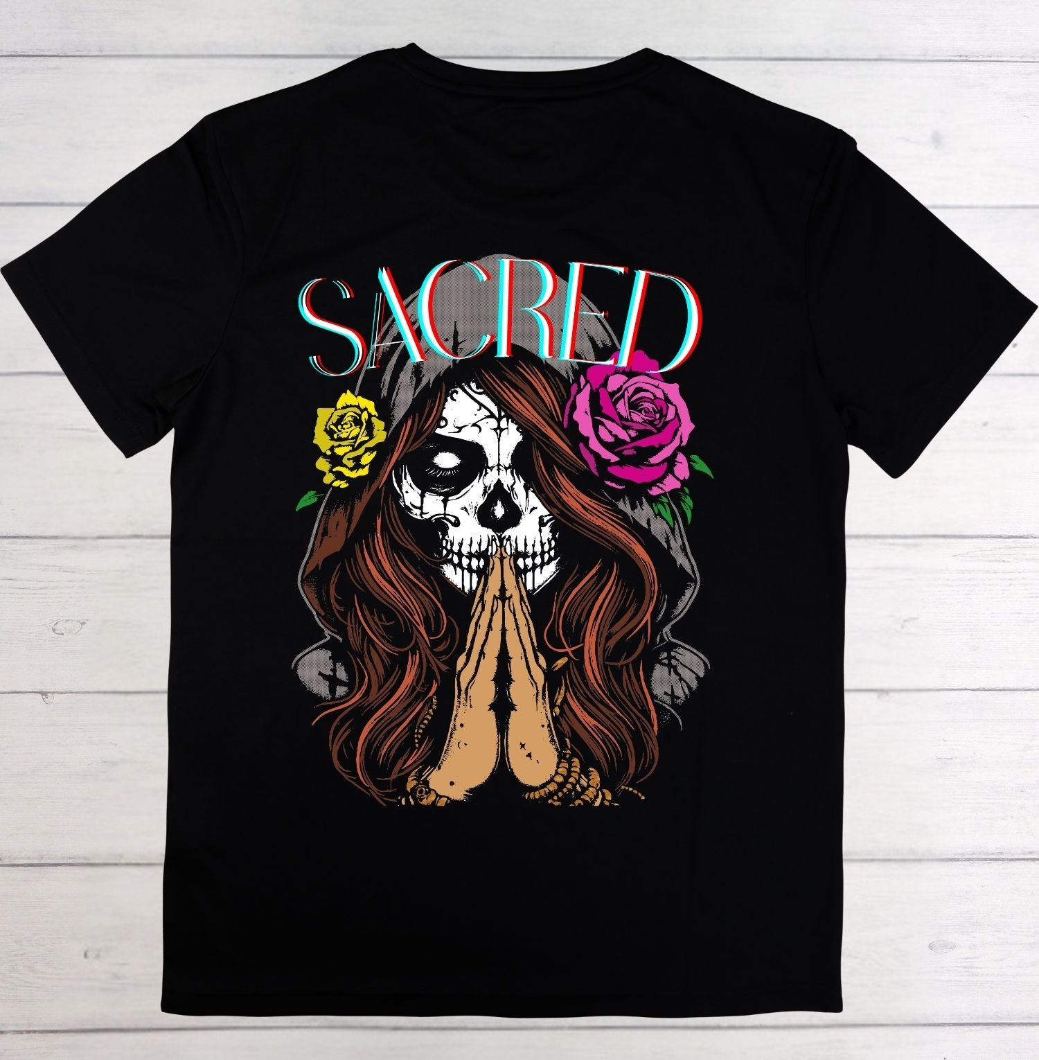 Black t-shirt with a graphic design of a skull with praying hands and flowers, labeled 'SACRED', on a wooden background.