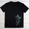 A black short-sleeve t-shirt laid flat on a white wood plank background, featuring a large graphic print of a giant footprint filled with a night forest scene, a Bigfoot figure, and a crescent moon.