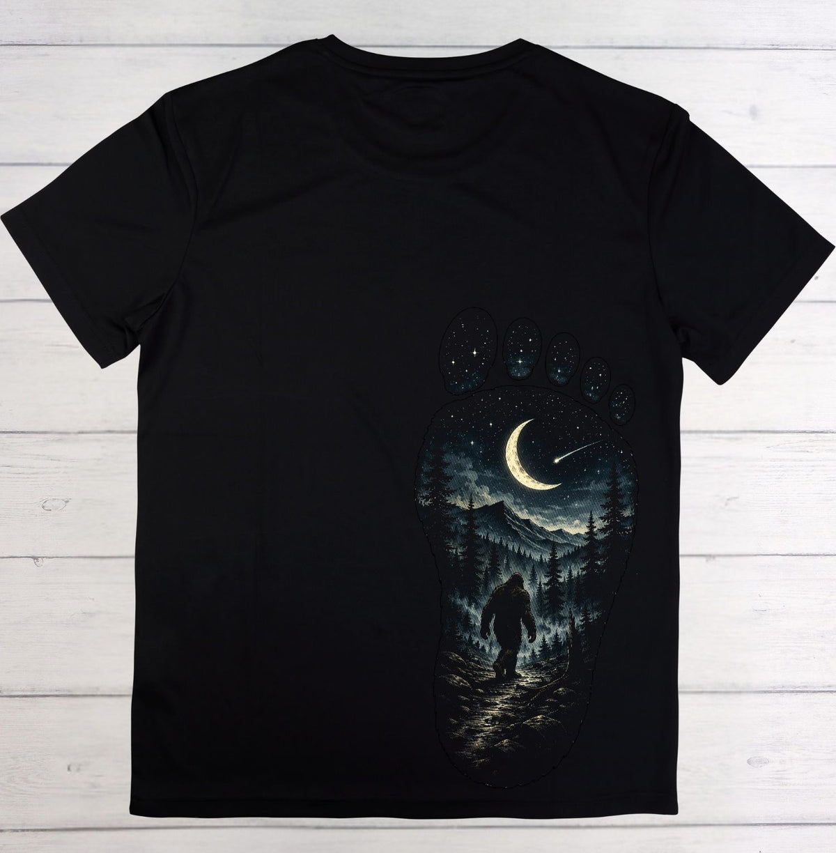 A black short-sleeve t-shirt laid flat on a white wood plank background, featuring a large graphic print of a giant footprint filled with a night forest scene, a Bigfoot figure, and a crescent moon.