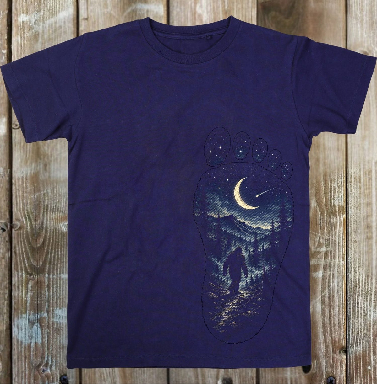A dark purple short-sleeve t-shirt with a graphic print of a Bigfoot silhouette inside a giant footprint, set against a rustic wooden plank background.