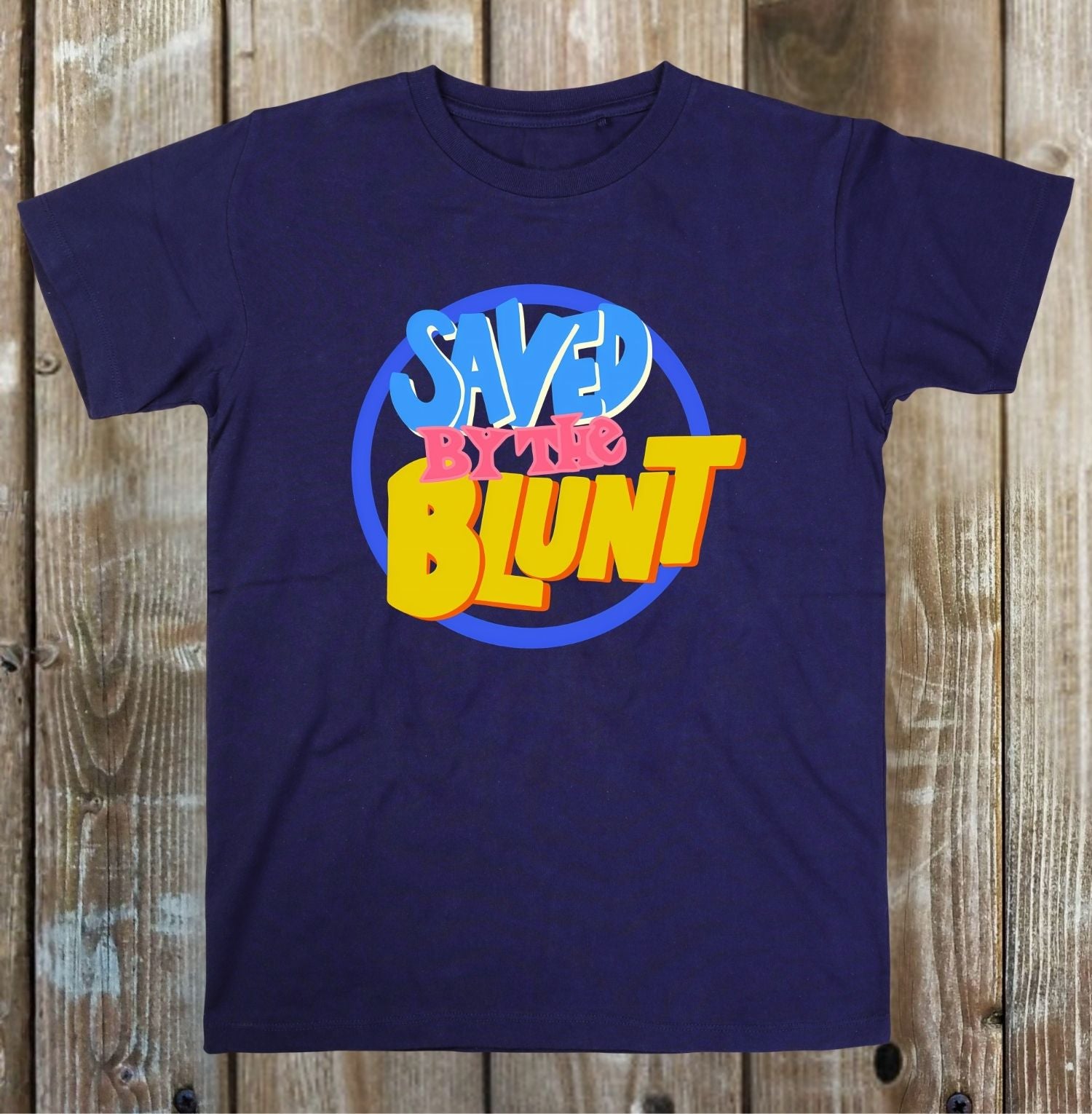 A navy blue short-sleeve t-shirt with a retro graphic reading 'SAVED BY THE BLUNT' in yellow, pink, and blue, laid flat on a rustic wooden plank background.