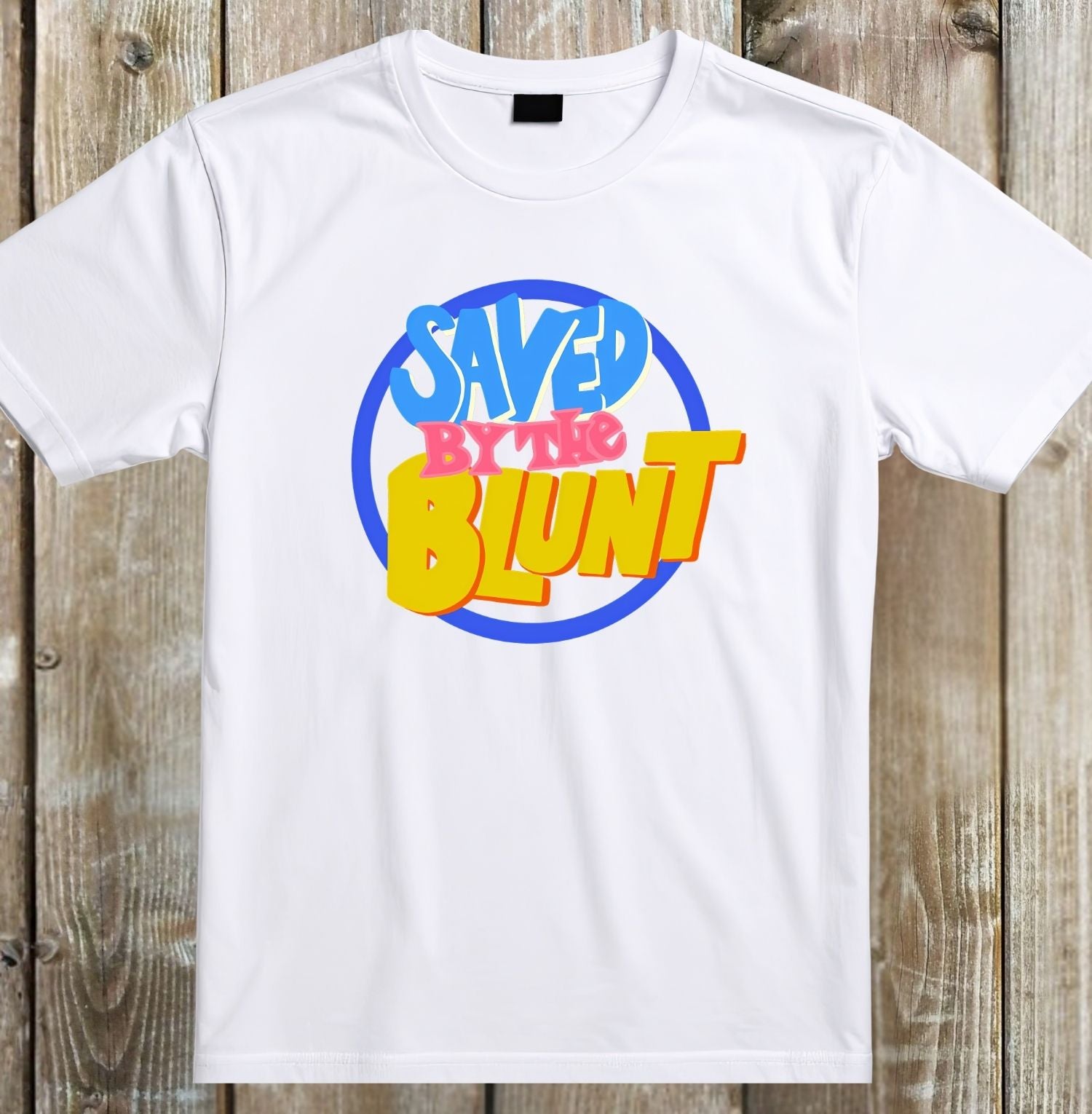 A white short-sleeve crewneck t-shirt with a colorful retro graphic on the chest reading 'SAVED BY THE BLUNT' in blue, pink, and yellow text inside a blue circle, displayed flat against a rustic wooden plank background.