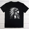 A black short-sleeve crewneck t-shirt featuring a detailed black and white graphic print of a woman in profile wearing a feathered headdress, laid flat on a light-colored wooden plank background.
