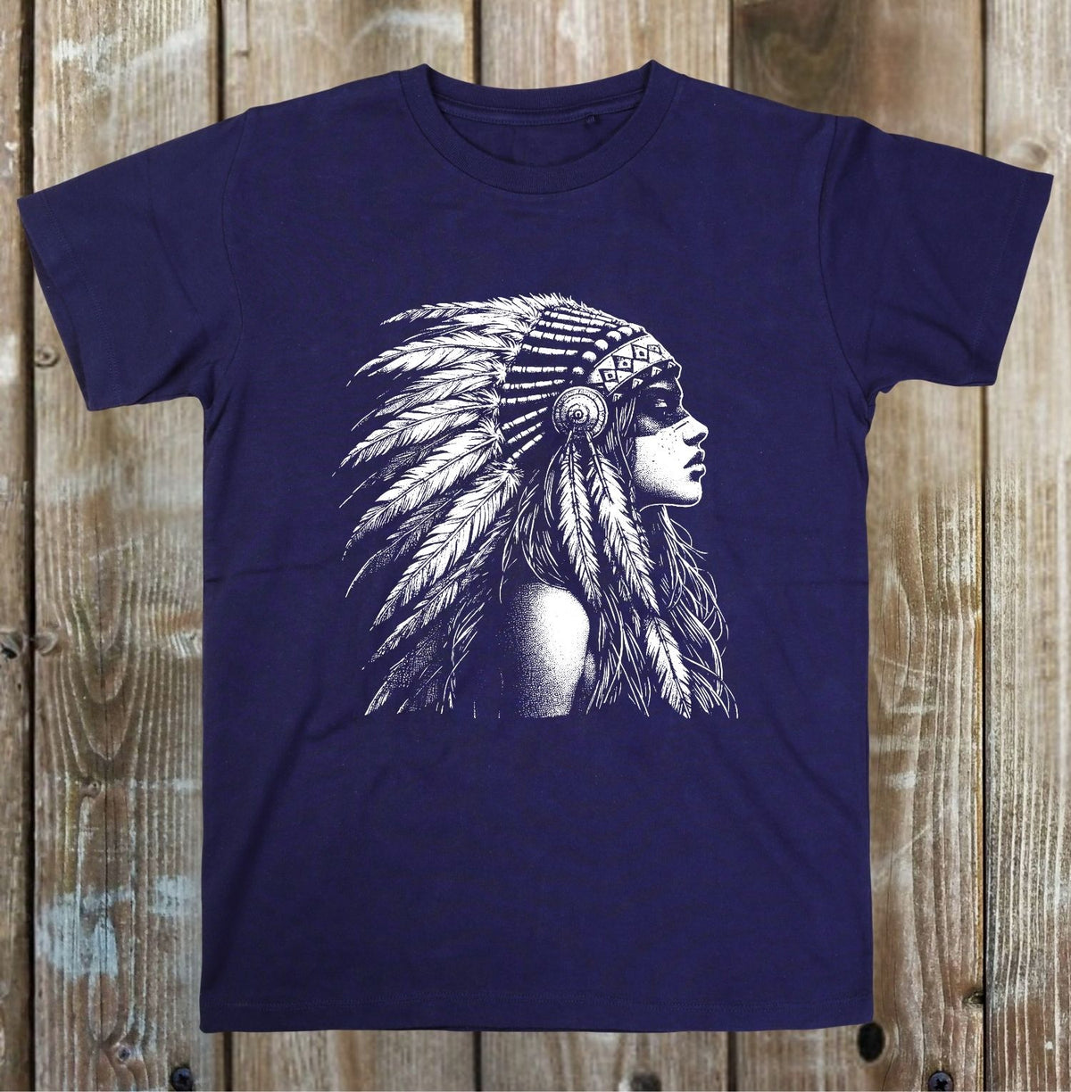 A navy blue short-sleeve t-shirt with a detailed white graphic of a woman in profile wearing a feathered headdress, displayed flat against a rustic wooden plank background.