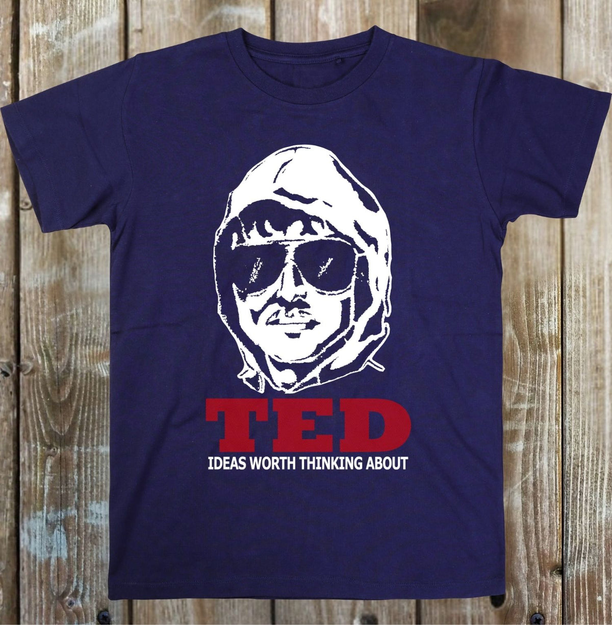 A navy blue short-sleeve t-shirt with a white graphic of a hooded figure wearing sunglasses and red text reading 'TED' with 'IDEAS WORTH THINKING ABOUT' underneath, displayed flat against a rustic wooden plank background.