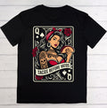 A black short-sleeve crewneck t-shirt laid flat on a white wood background, featuring a large graphic print of a Queen playing card illustration with a woman holding a taco and the text 'TACOS BEFORE VATOS'.