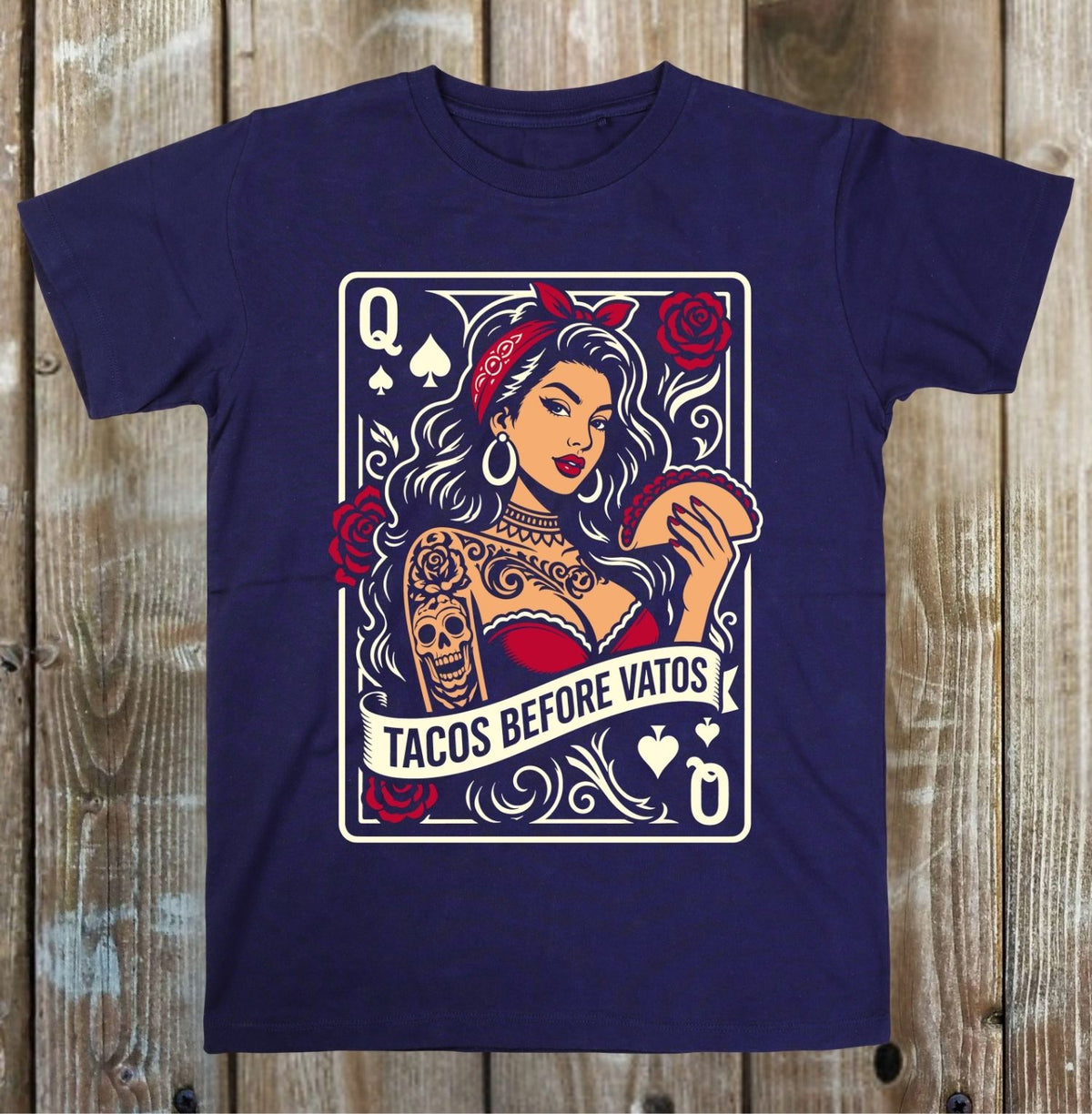 A navy blue short-sleeve t-shirt with a graphic print of a playing card design featuring a woman with tattoos, red roses, and the text 'TACOS BEFORE VATOS', displayed flat against a rustic wooden plank background.