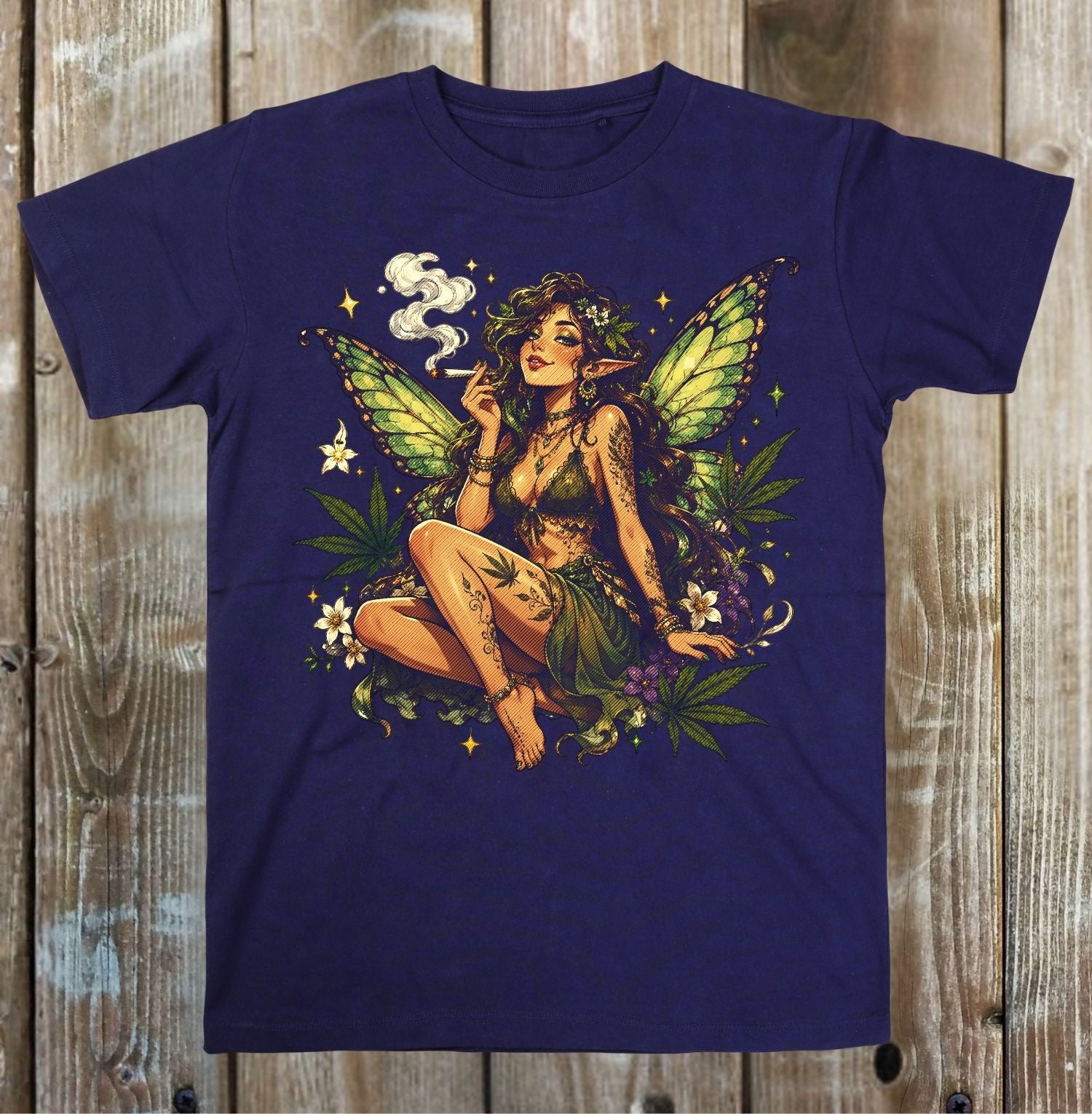 A navy blue short-sleeve t-shirt with a detailed graphic of a female fairy smoking a joint, surrounded by green cannabis leaves and small white flowers, displayed flat against a rustic wooden plank background.