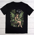 A black short-sleeve crewneck t-shirt laid flat on a white wood plank background, featuring a large graphic print of a female fairy with green wings, tattoos, and cannabis leaves and small white flowers.