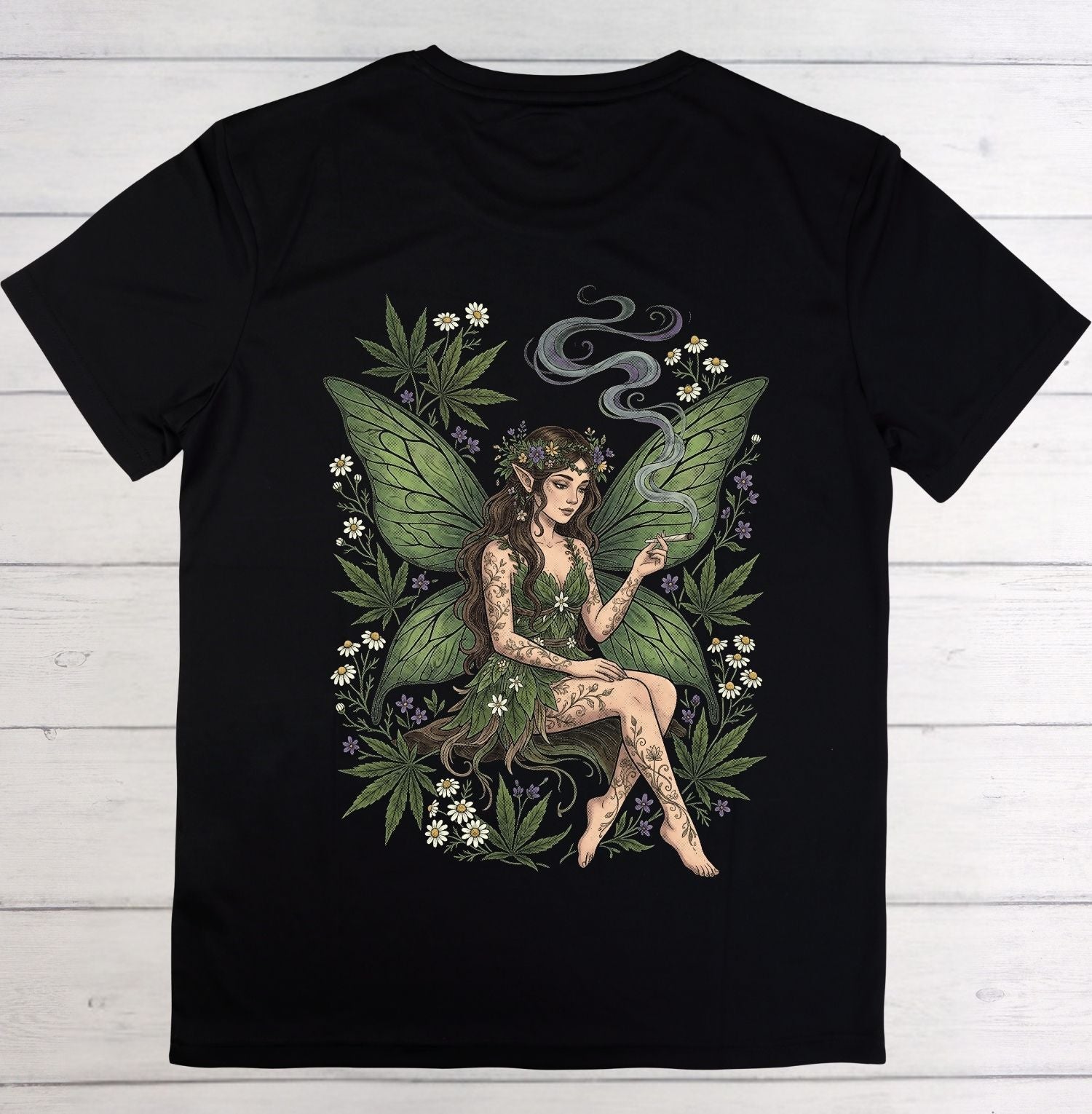 A black short-sleeve crewneck t-shirt laid flat on a white wood plank background, featuring a large graphic print of a female fairy with green wings, tattoos, and cannabis leaves and small white flowers.