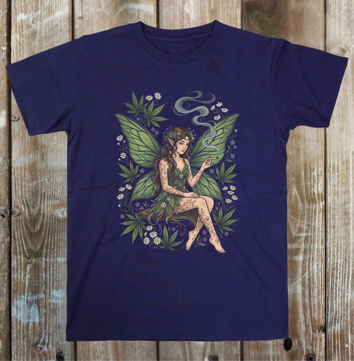 A navy blue short-sleeve t-shirt with a detailed graphic of a fairy sitting among green leaves and small white flowers, displayed flat against a rustic wooden plank background.