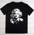 A black short-sleeve t-shirt laid flat on a white wood plank background, featuring a large black-and-white graphic print of Marilyn Monroe holding a rose on the front.