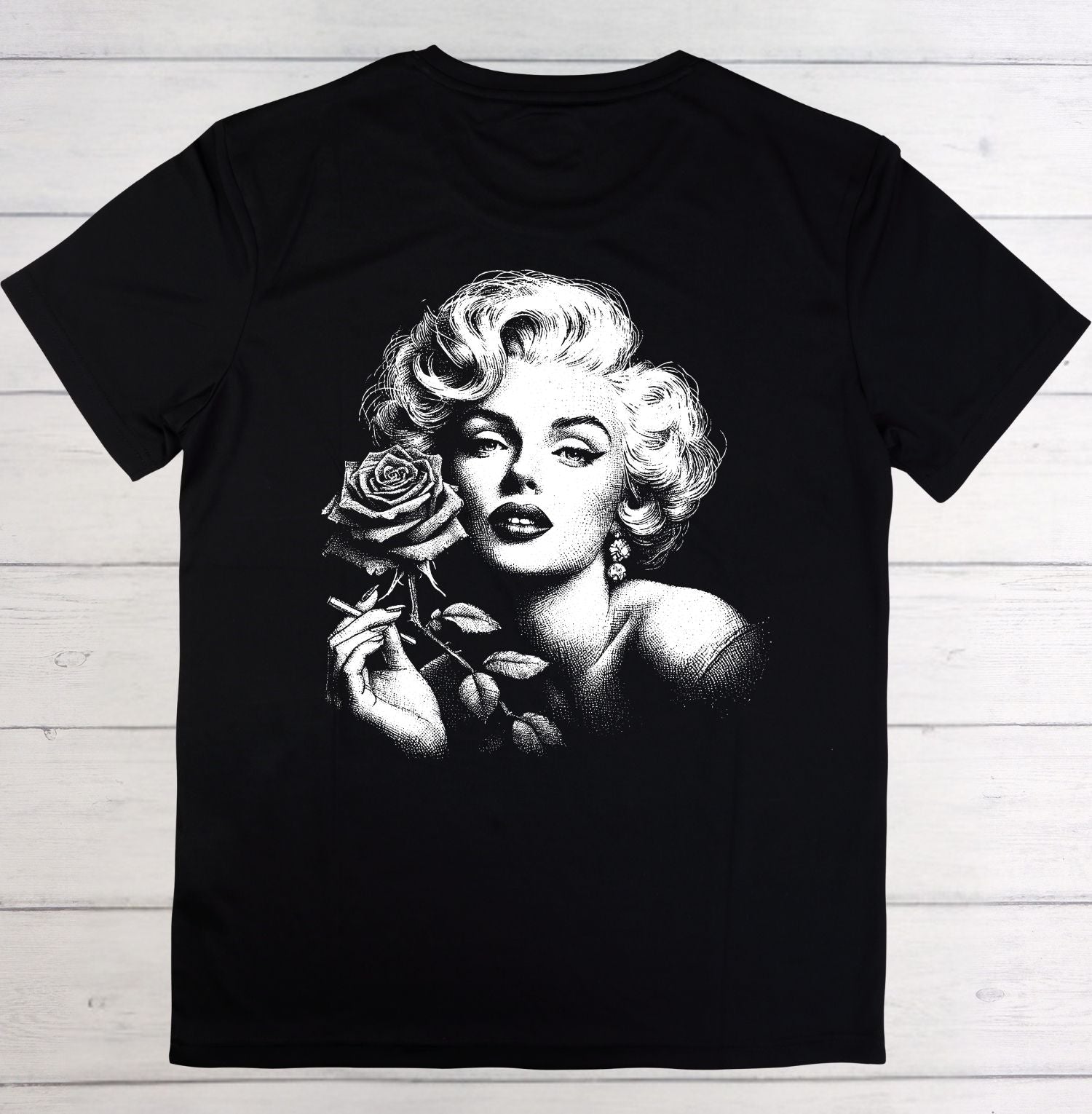 A black short-sleeve t-shirt laid flat on a white wood plank background, featuring a large black-and-white graphic print of Marilyn Monroe holding a rose on the front.