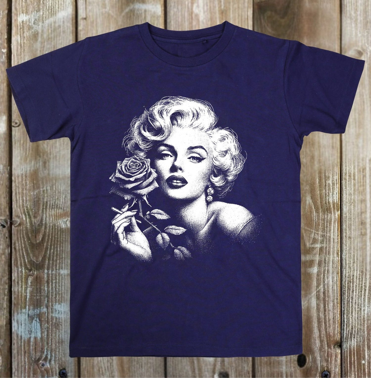 A navy blue short-sleeve t-shirt featuring a large white graphic print of Marilyn Monroe holding a rose, displayed flat against a rustic wooden plank background.