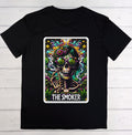 A black short-sleeve crewneck t-shirt laid flat on a white wood plank background, featuring a large colorful graphic print of a tarot card labeled 'THE SMOKER' with a skull, skeleton hand, and green leaf motifs.