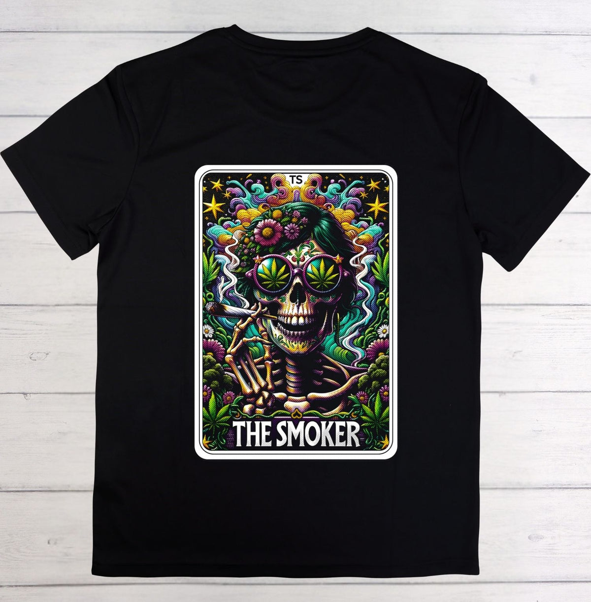 A black short-sleeve crewneck t-shirt laid flat on a white wood plank background, featuring a large colorful graphic print of a tarot card labeled 'THE SMOKER' with a skull, skeleton hand, and green leaf motifs.