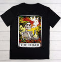 A black short-sleeve crewneck t-shirt laid flat on a white wood background, featuring a large graphic print of a tarot card styled as 'The Toker' with a skeleton, cannabis leaves, and a bottle of beer.