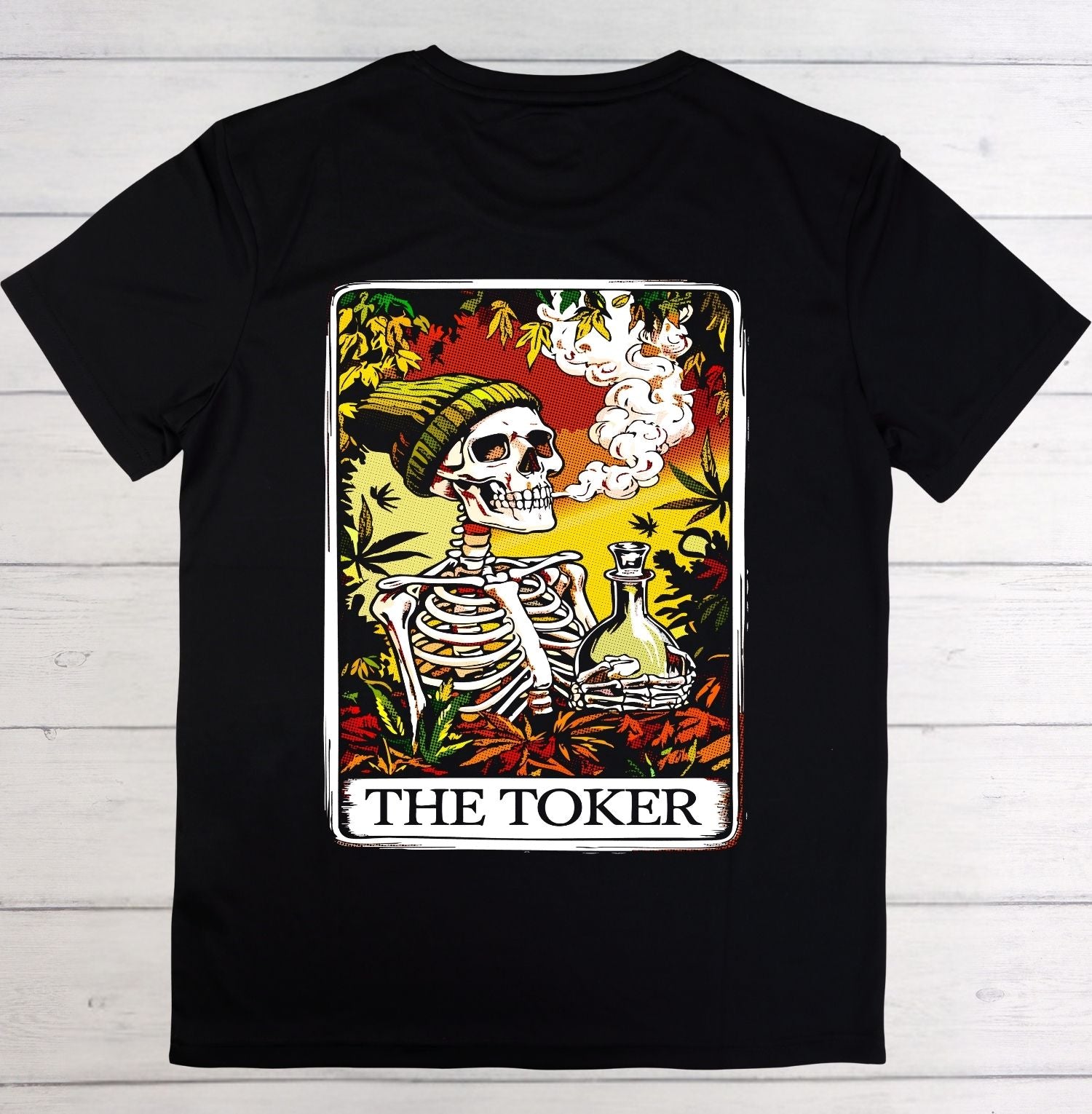 A black short-sleeve crewneck t-shirt laid flat on a white wood background, featuring a large graphic print of a tarot card styled as 'The Toker' with a skeleton, cannabis leaves, and a bottle of beer.