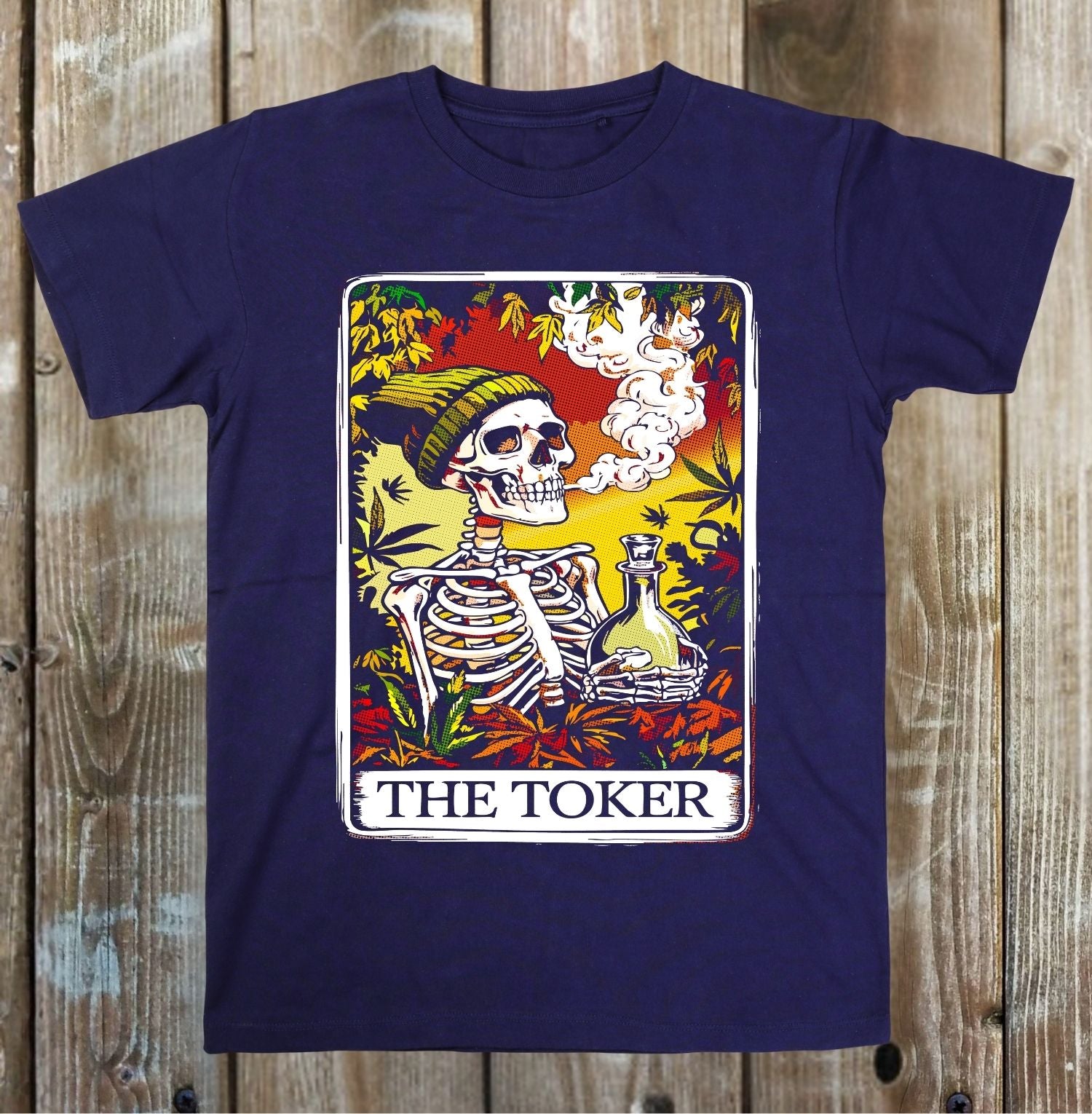 A navy blue short-sleeve t-shirt with a colorful graphic print of a tarot card titled 'THE TOKER', featuring a skeleton, a beanie, and a bong, laid flat on a rustic wooden plank background.